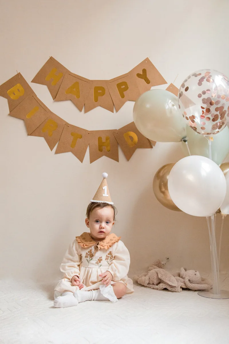 Baby in a party hat on a neutral set with bunting and balloons