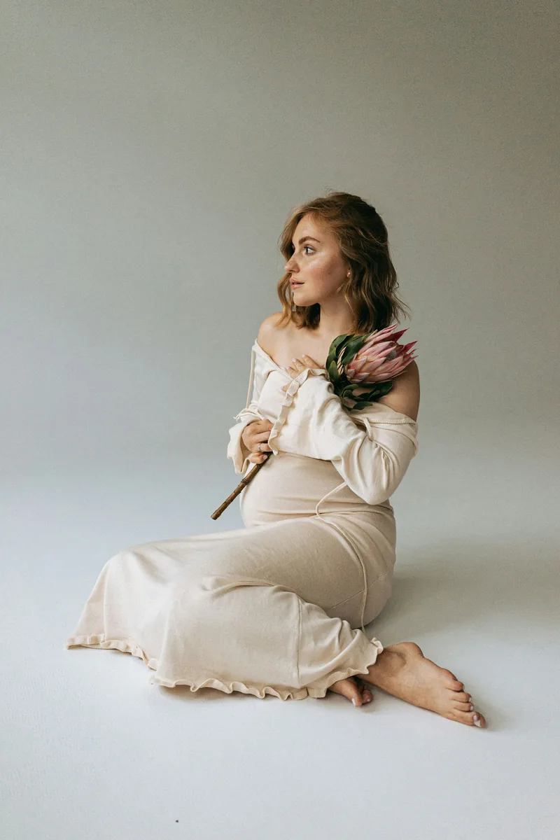 Expecting mother seated on the studio floor in a cream gown, holding a single flower