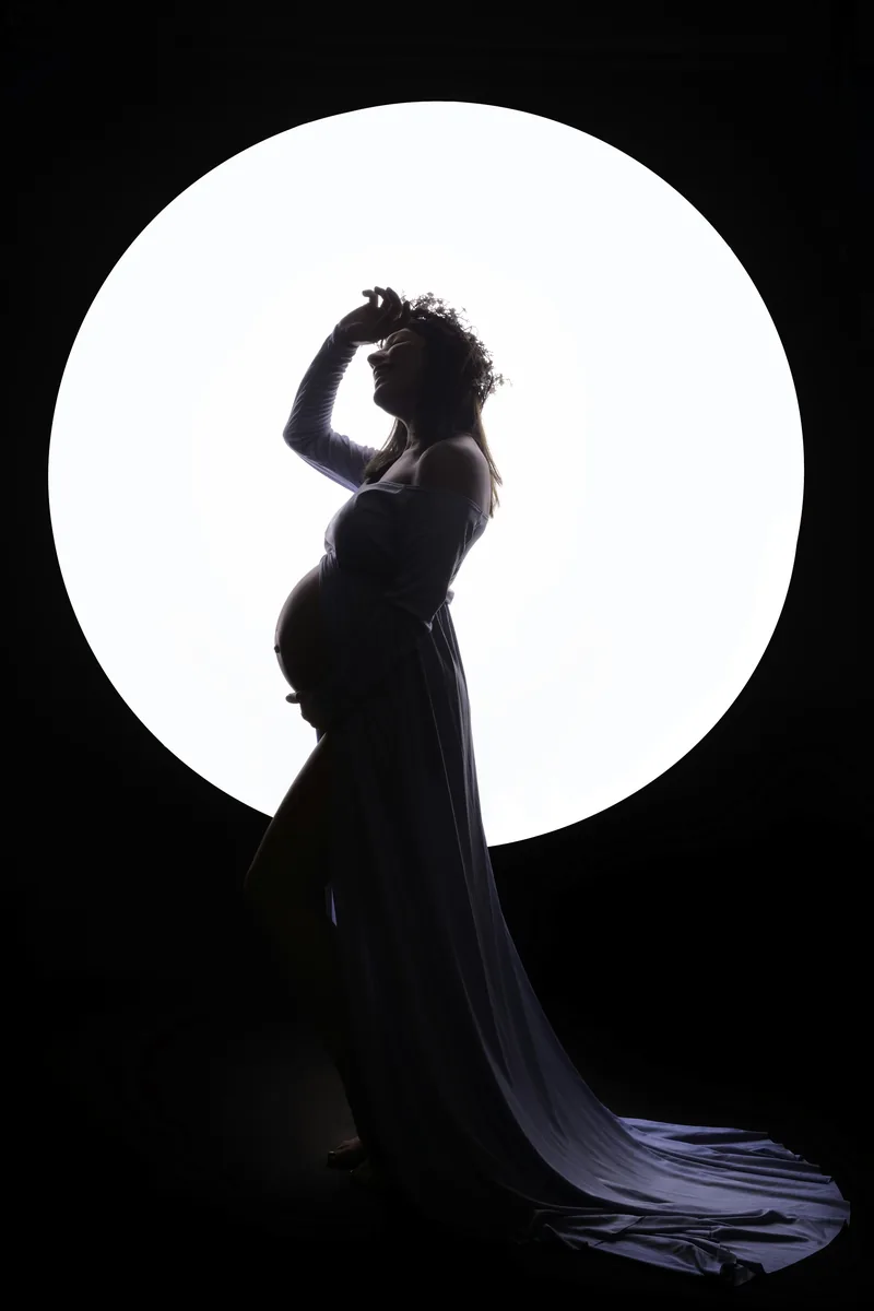 Maternity silhouette against a glowing circular backdrop, gown trailing on the floor