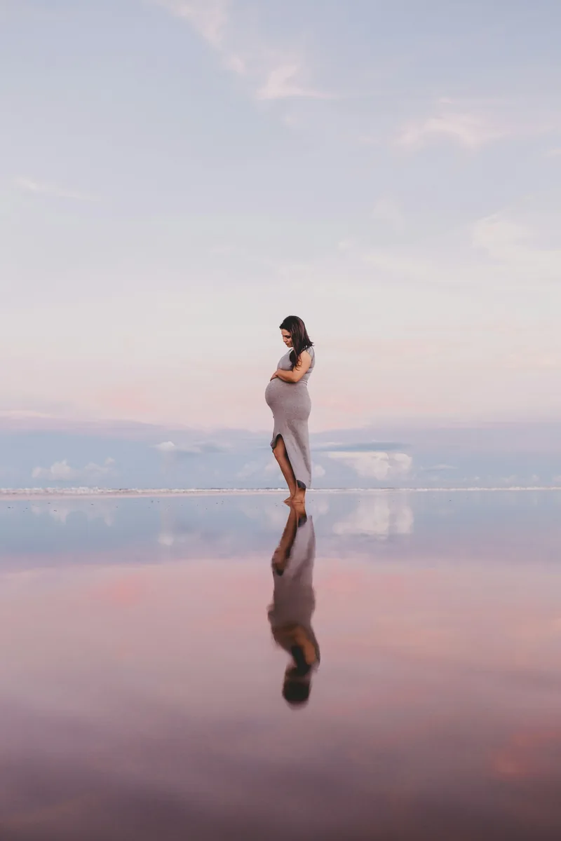 Expecting mother standing on a mirrored shore under a soft pink sky