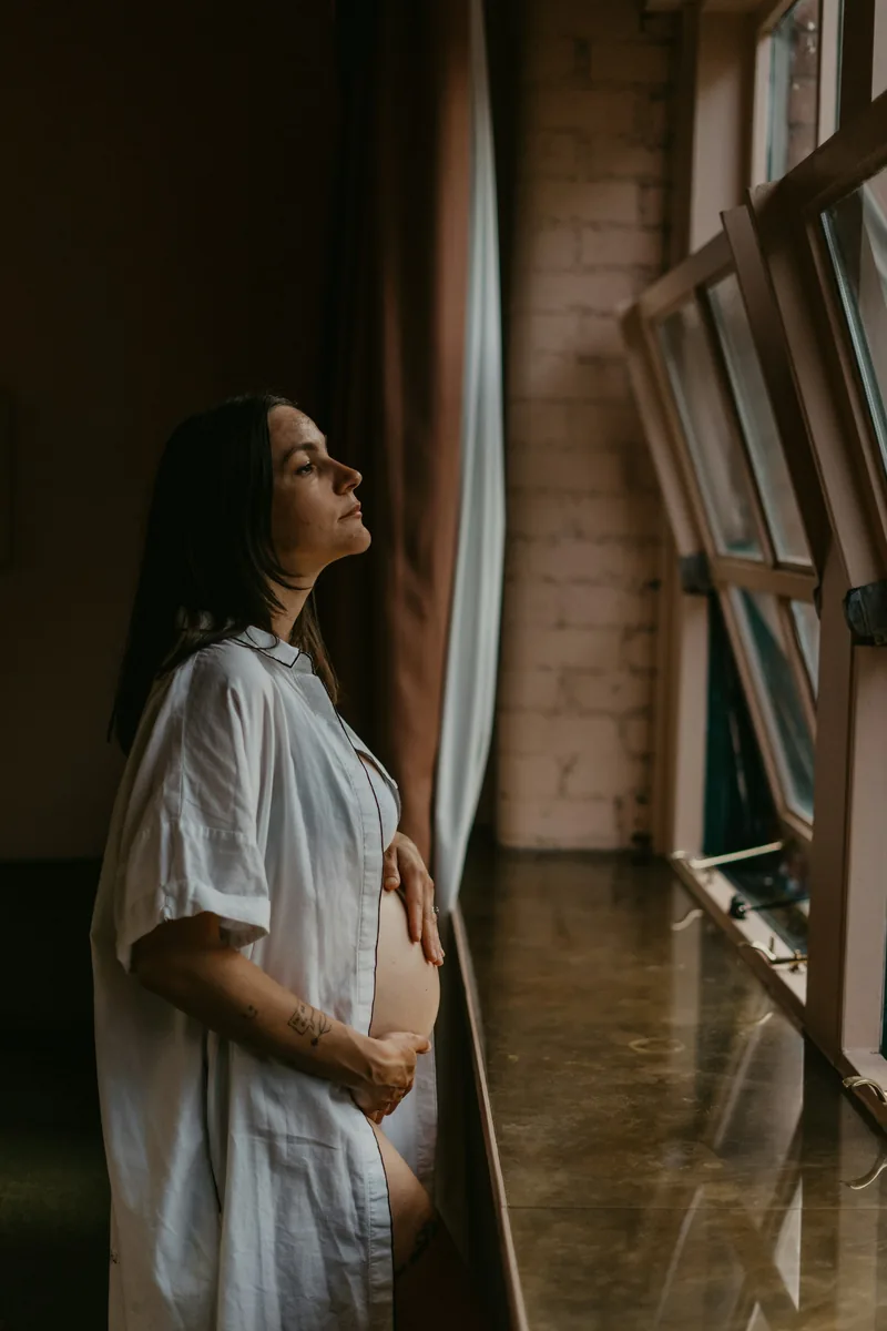 Expecting mother by tall windows in soft, moody daylight