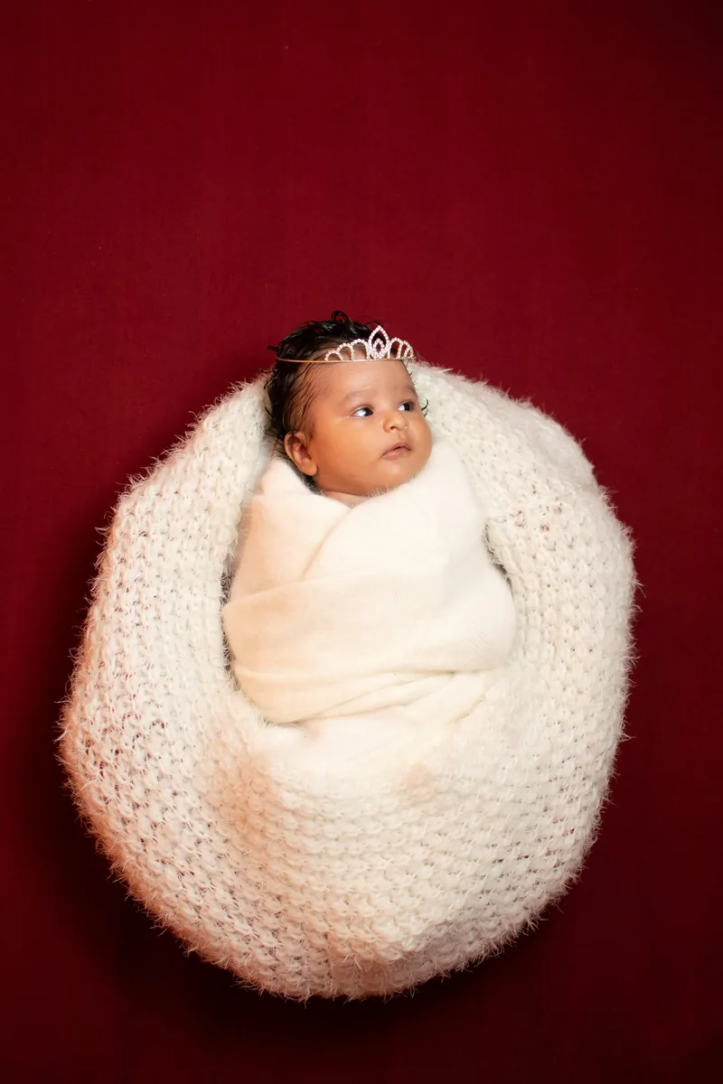 Newborn with a tiny tiara in a cream knit nest against deep red